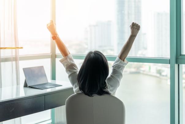 A woman sitting at an office desk raises her arms in the air with excitement.