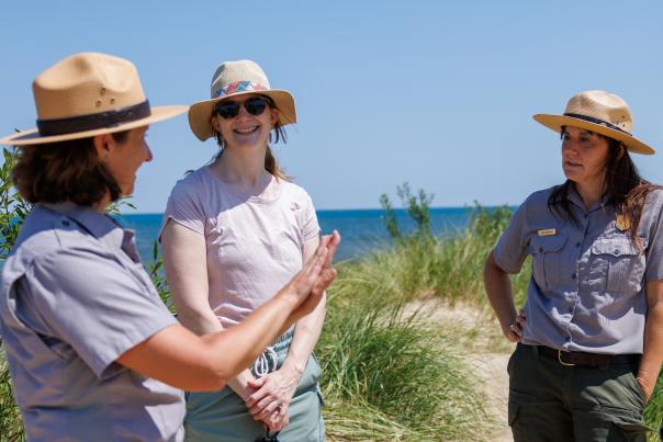 A woman in a straw hat, sunglasses, and casual outdoor clothing stands on a sandy path near Lake Michigan, smiling as she talks with two park rangers. The rangers wear gray uniform shirts, dark green pants, and wide-brimmed ranger hats. One ranger gestures with her hand while speaking, while the other listens with a serious expression. Behind them, tall beach grass and the deep blue lake stretch toward the horizon under a clear sky.