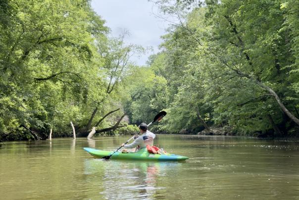 Person kayaking down the calm Flint River surrounded by lush green trees and peaceful wooded scenery near Huntsville, Alabama.