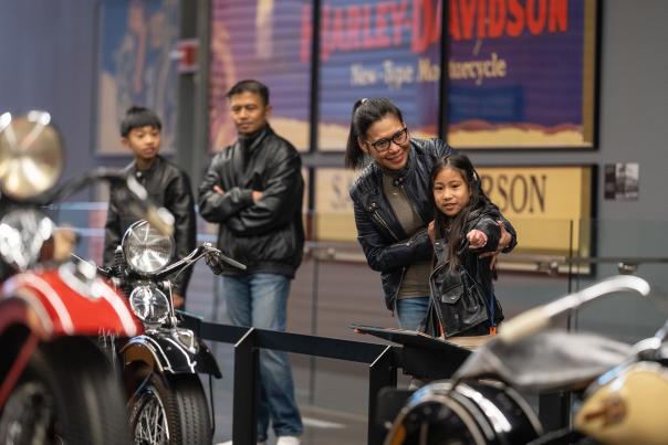 A family of four, all dressed in black leather jackets, visits the Harley-Davidson Museum. A smiling woman and a young girl in the foreground point at a vintage motorcycle display, while two other family members stand behind them observing.