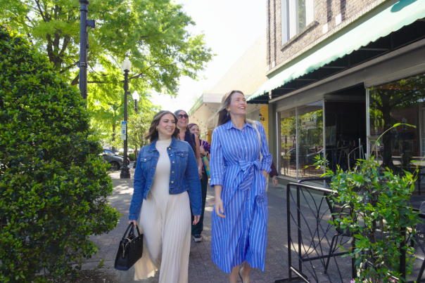 Two women walking along a tree-lined sidewalk in downtown Fayetteville, NC, passing local shops and outdoor dining.