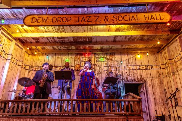 A jazz ensemble performs on the rustic wooden stage at Dew Drop Jazz Hall in Mandeville beneath the historic “Dew Drop Jazz & Social Hall” sign and colorful string lights.