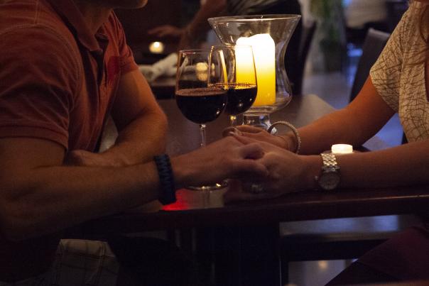 A couple hold hands while enjoying glasses of red wine at table.