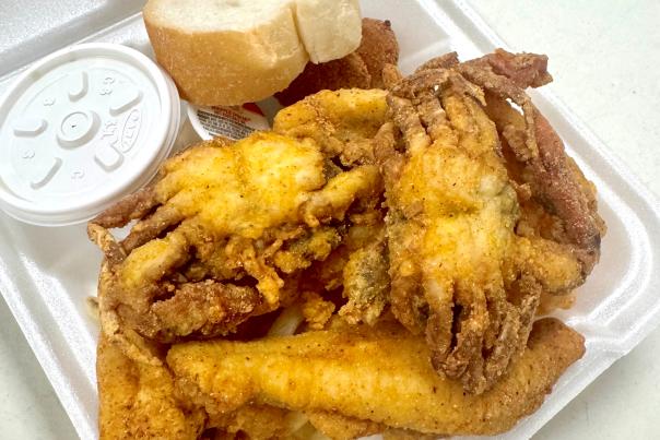 Fried catfish, soft shelled crab and fries with a small white cup of dipping sauce and a piece of bread, piled in a Styrofoam box.