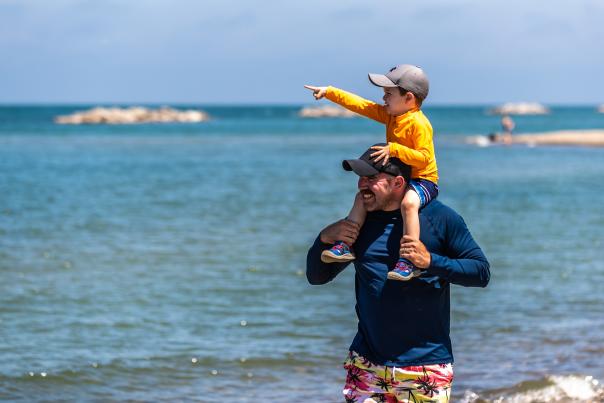 A young boy on a father's shoulders walking on a beach. The young boy is pointing out at an ocean
