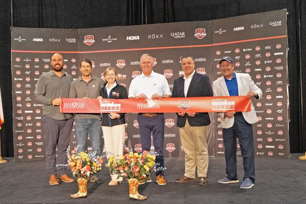 Six people on a stage hold a thick red ribbon that reads, "MEMORIAL HERMANN," "MEMORIAL HERMANN IRONMAN TEXAS," and "IRONMAN PRO SERIES" in repeat across it. In the middle are Linda Nelson and Brad Bailey. On the far right is Nick Wolda.