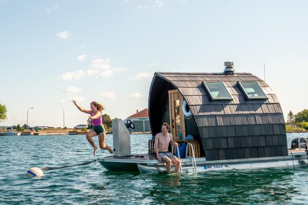 Two people jumping off of the Sisu & Loyly floating sauna in the Grand Marais harbor