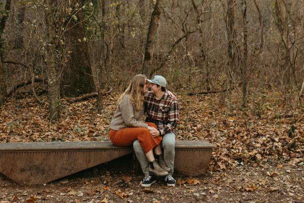 A queer couple sits on a metal bench in Coler Mountain Bike Preserve, surrounded by bare trees and fallen autumn leaves. One wears an orange skirt and gray sweater, leaning toward the other in a plaid shirt and blue cap. The forested setting reflects the preserve’s natural beauty and inclusive spirit.
