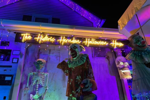 A house decorated in purple and orange glowing light with a skeleton and scary looking clown with jagged teeth in foreground. A lit up sign reads in the back that says The Historic Herndon Halloween House.