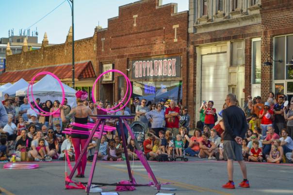 Hoop dancer with neon pink hoops twirls 8 hoops around her body in front of an audience seated on the street.