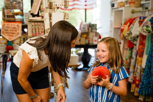 The camera focuses on a mom and kindergarten-age daughter. The mom (in a white blouse and navy shorts) leans down to talk with her child (wearing a blue and white striped dress). The girl is laughing and holding an apple-shaped plush to her chest. Behind them is a local boutique featuring school supplies, trendy dresses, and more.