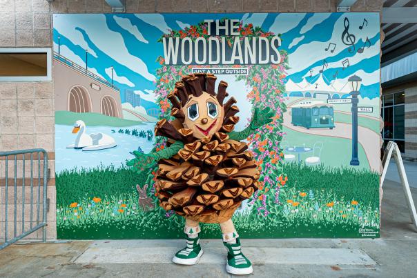 Puffy The Pinecone Poses in front of Visit The Woodlands Mural