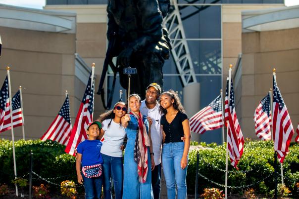 Family posing together in front of a large military statue with American flags displayed outside a museum building