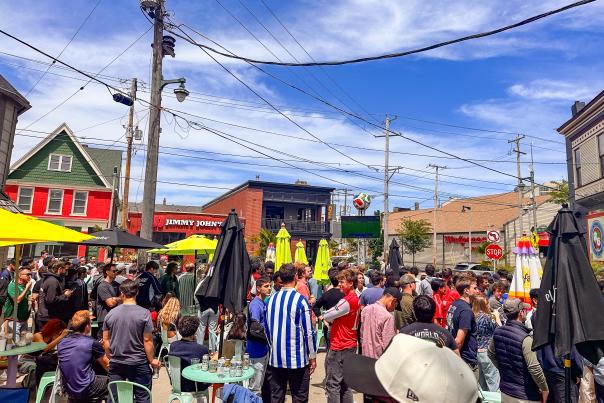 Soccer fans pack the patio and surrounding area outside Nomad World Pub on Brady Street in Milwaukee, watching a match on a large outdoor screen during a sunny afternoon.