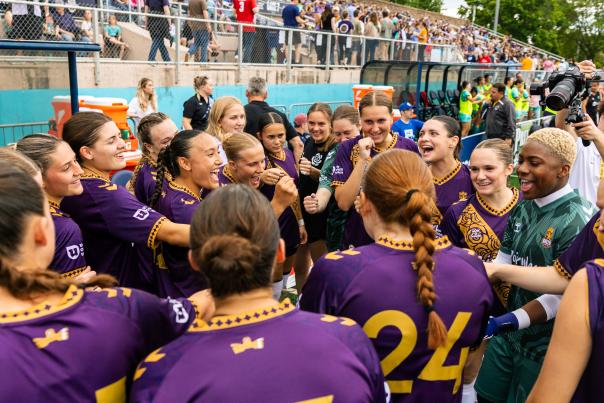 A soccer team in purple jerseys gathers in a tight pregame huddle along the sideline. Behind the group, a crowded set of bleachers lines the field, separated by a chain-link fence.