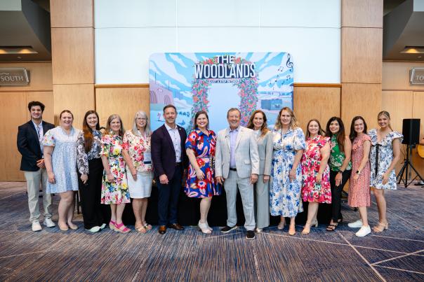 Fourteen people smile at the camera in front of a colorful Visit The Woodlands backdrop. From left: Ryan Greenwood, Vargo Williams, Rachael Varner, Ashley Fenner, Julie Quinn, Adam Lamb, Elizabeth Eddins, Nick Wolda, Cindy Heiser, Josie Lewis, Amber Parsley, Carla Twidell, Andrea Day, and Brooklyn Newell.