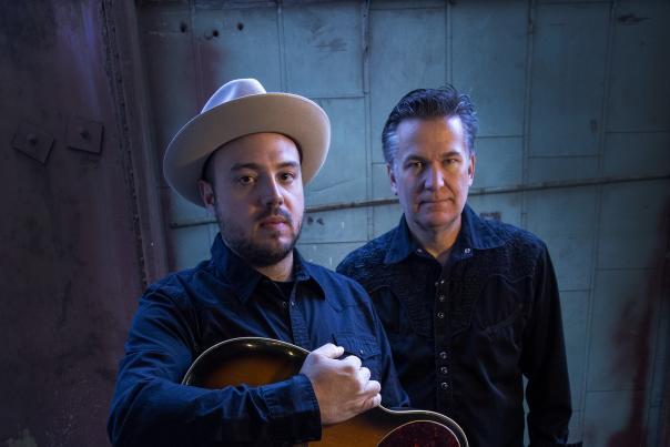 Two men look up at the camera. Both are in black. The one in the front holds a guitar and wears a hat.