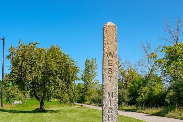 Concrete, pyramidal shaped West Michigan Pike Marker rises above green grass. Blue sky, cement bike path and green trees are in background.