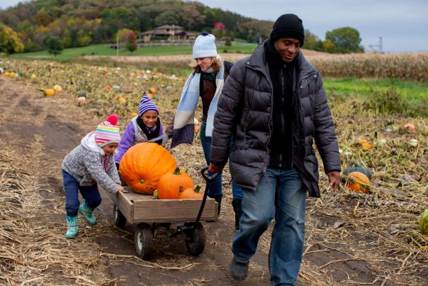 A bi-racial family at Treinen Farms. The dad is pulling a wagon with three large pumpkins while his daughters and wife follow behind him.