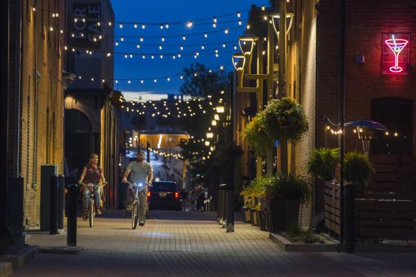 A couple rides bikes in downtown underneath the alley lights