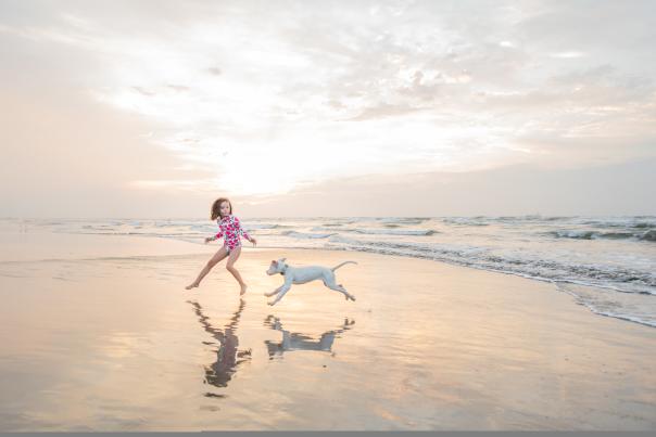 A girl in a pink rash guard runs on the beach with a white dog in the early morning.