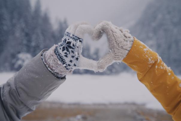 Two gloved hands form a heart shape against a snowy landscape, with evergreen trees in the background and soft snowfall.