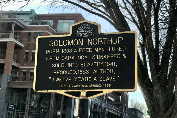 Solomon Northrup historical marker sign facing south on Broadway with a bare tree and building behind it.