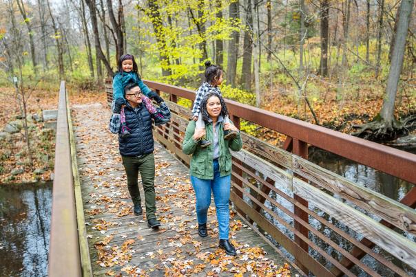 A family of four walks along a leaf covered bridge in For-Mar Arboretum and Nature Preserve