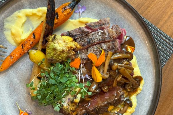 The camera focuses on an expertly plated meal at The Waterway Marriott. It features a nearly rare steak, cut into strips, and it's topped with a BBQ-like sauce, long and skinny mushrooms, orange flower petals, and sprouts. It is served with several cooked carrots (yellow and orange) and cooked cauliflower, and it's served on a thick yellow saffron sauce.