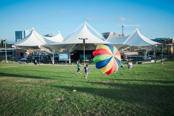 A daytime photo of The Pavilion tents. Two young families play with colorful parachutes on the lawn.