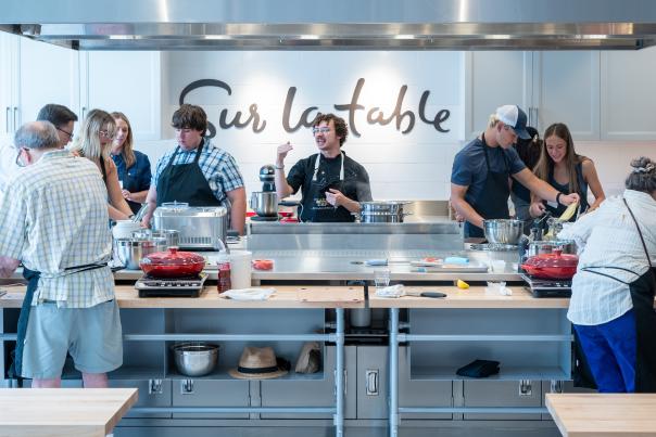 A young chef with curly brown hair, round glasses, and a black denim apron instructs a cooking class (at least 10 men and women of various ages and backgrounds are visible). Various pots, pans, hot plates, and other cooking utensils are spread out on the counter before them. On the wall in the back is "Sur la table" written in large, black, cursive.