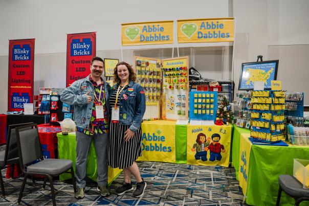 Married couple and owners of Abbie Dabbles bricks and minifigs accessories, Abbie and E.J., pose with their booth at BAMCOM, hosted at The Woodlands Waterway Marriott. E.J. has a curly mustache and is wearing a bright, multicolored shirt with a denim jacket bearing various patches he created. Abbie is wearing a black-and-white striped dress with a denim jacket like E.J.'s.