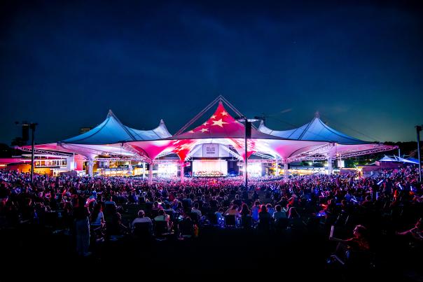 A wide-angle view of The Pavilion during the 2024 Star-Spangled Salute. The tents are lit up red, white, and blue with projections of stars across the red. The Houston Symphony is barely visible on stage, but every seat is packed from the front row to the back of the lawn. The sky is dark navy.