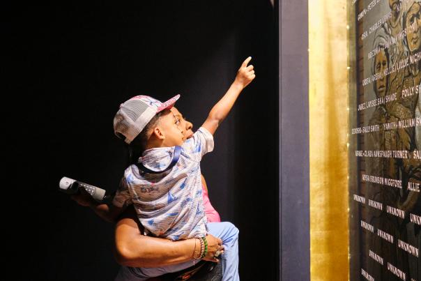 African American man holding a young child in front of the plaque with engraved names of the Clotilda, the Last American Slave Ship
