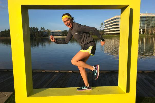 A woman in athletic clothes poses as if she's running inside a public art piece. The sculpture is of a large yellow square, like a window without the glass, overlooking Lake Woodlands at Hughes Landing.