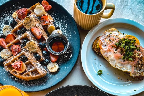 Overhead view of a colorful brunch spread at The Artist Village Café in Santa Ana with fruit-topped waffles, French toast with strawberries and powdered sugar, avocado toast with eggs, coffee, and orange juice.