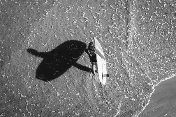 Black and white aerial image of a woman holding a surf board in the shallow water. Her shadow stretches out to the left.