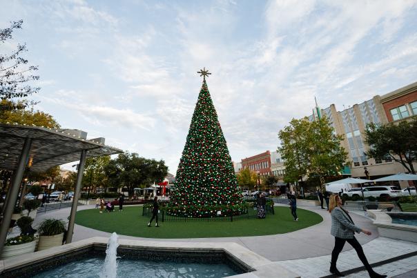 The Christmas Tree at Market Street is 70 feet tall, ringed with lights and decked with ornaments in red and green. A golden eight-point star rests at the top. The photo is taken from in front of Sixty Vines, facing the tree, a fountain, and the Market Street marquee.