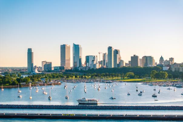 A wide view of Milwaukee’s lakefront skyline on a clear day, with dozens of sailboats anchored in a calm marina and a tour boat cruising along the breakwater in the foreground. Green parkland lines the waterfront beneath a cluster of downtown high-rise buildings and glass towers.