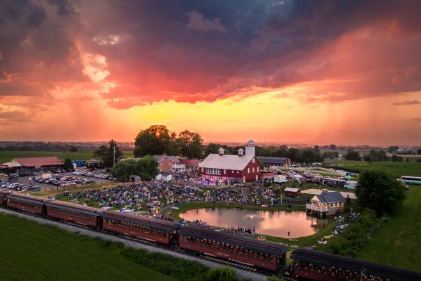 Aerial view of Paradise Station in Lancaster with a large crowd of people on grass in front of a large red barn with a dark sunset overhead