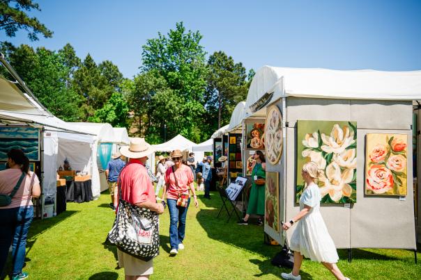 A wide-angle view of The Woodlands Waterway Arts Festival at Town Green Park. Visible is a winding row of white pop-up tents layered with colorful paintings, photos, and other works of fine art. Visitors mill about in and among the tents, admiring the displays. Behind everything are bright green trees and a bright blue sky.