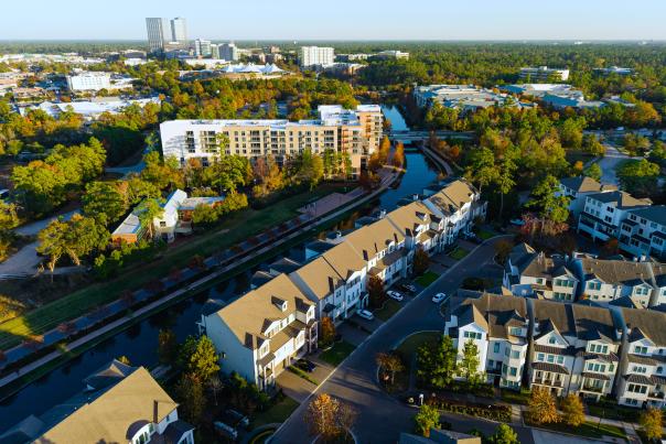 An aerial drone view of The Woodlands during late afternoon. The camera hovers over residences along the south/west arm of The Waterway, facing The Pavilion and The Woodlands Towers.