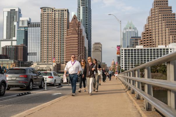 A group of people wearing SXSW badges walk across the Congress Avenue Bridge with the Austin skyline in the background.