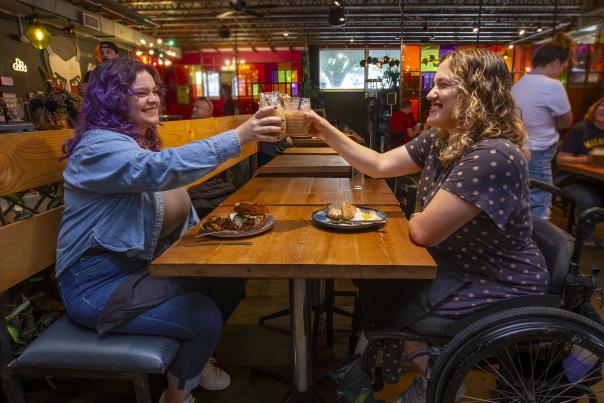 A woman with purple hair clinks glasses with a smiling woman in a wheelchair above plates full of food at the People's Kitchen.