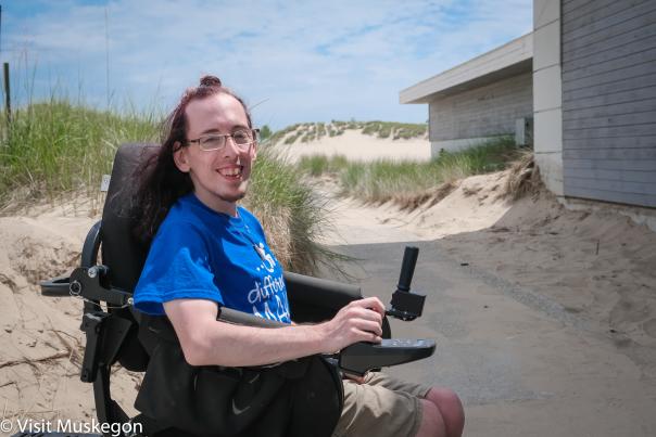 Person in a wheelchair smiling on a sandy beach near grass-covered dunes, wearing a blue t-shirt