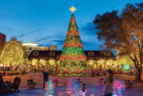 The Market Street Christmas Tree is twice as tall as the building behind it, wrapped in golden Christmas lights with alternating stripes of red and green lights. The trees around it are also wrapped in golden lights. Families play in the splash pad near the camera.