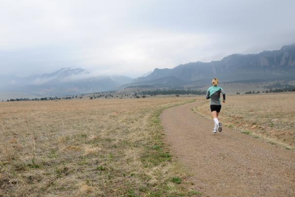 A woman jogs along a winding gravel trail through open grasslands beneath misty skies and the Flatirons in Boulder, Colorado.