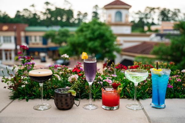 Six colorful drinks are lined up on the balcony at Hyatt Centric's rooftop pool. On the far left, an espresso martini, then a mule with lime and sprig of rosemary, then a tall purple cocktail, then a bright red drink with a strawberry and jalapeno slice on the rim, then a cucumber martini, then a tall blue drink. In the background is an (out of focus) overview of Market Street.