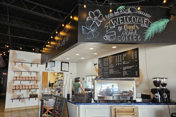 Interior of Angel's Island Coffee featuring a bright coffee counter, espresso equipment, menu boards, coffee bean grinders, and shelves stocked with bags of coffee. A large hand-drawn chalk mural reading "Welcome to Angel's Island Coffee" with tropical flowers and palm leaves decorates the space, while string lights hang from the open ceiling, creating a warm and inviting atmosphere.