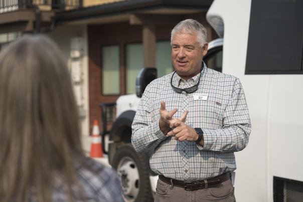 A man in a plaid shirt gestures while speaking to a woman, with a vehicle and buildings in the background. The scene suggests a guided experience.
